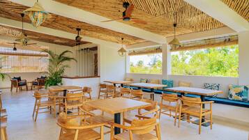 Un espace intérieur spacieux, doté de tables et de chaises en bois, d’un ventilateur de plafond et de grandes fenêtres offrant une vue sur la verdure environnante.