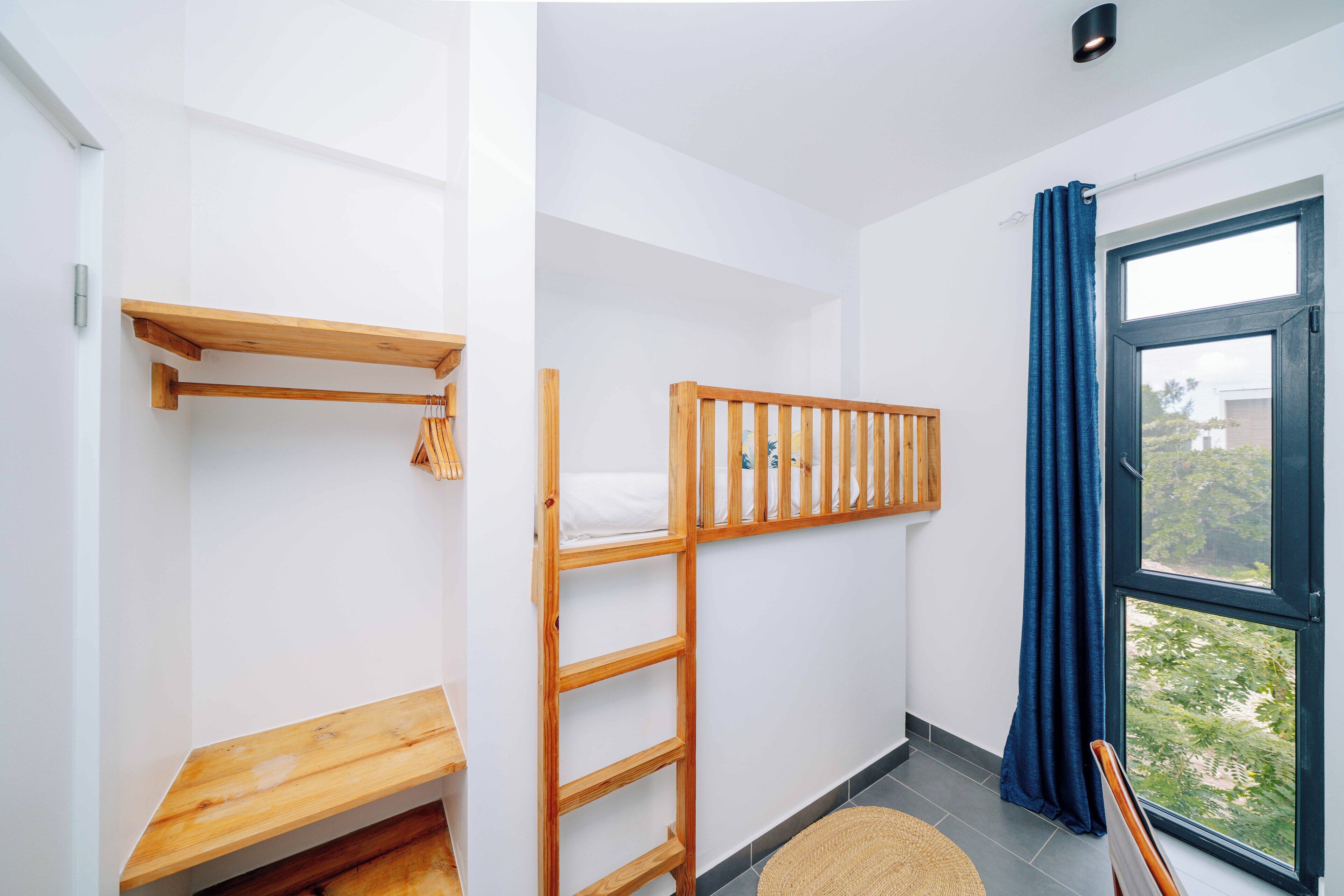 A room with a bunk bed, a wooden shelf, a ladder, a small table, and a window with curtains.