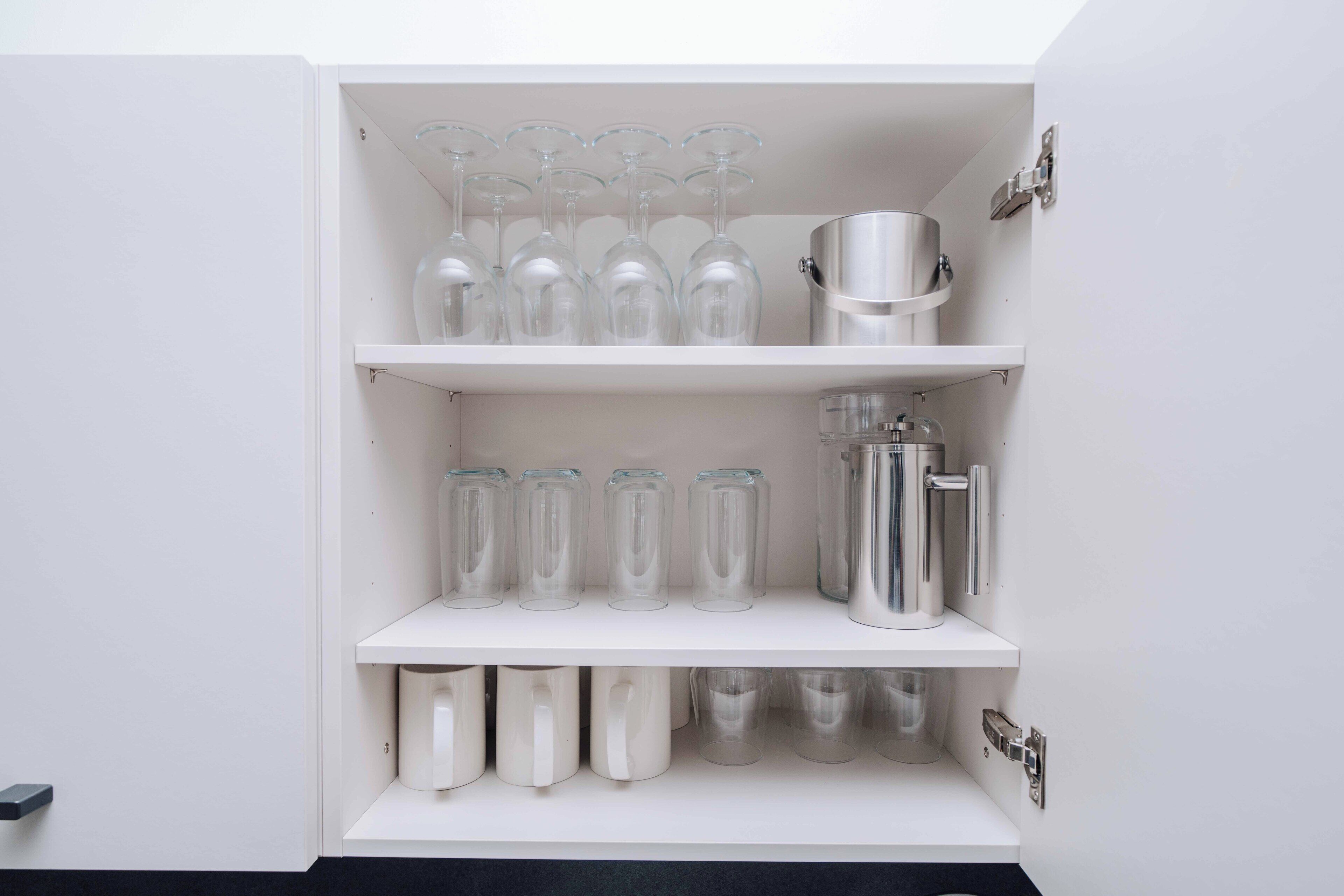 Um armário branco com prateleiras contendo copos de vidro transparentes, potes de vidro e recipientes de aço inoxidável.