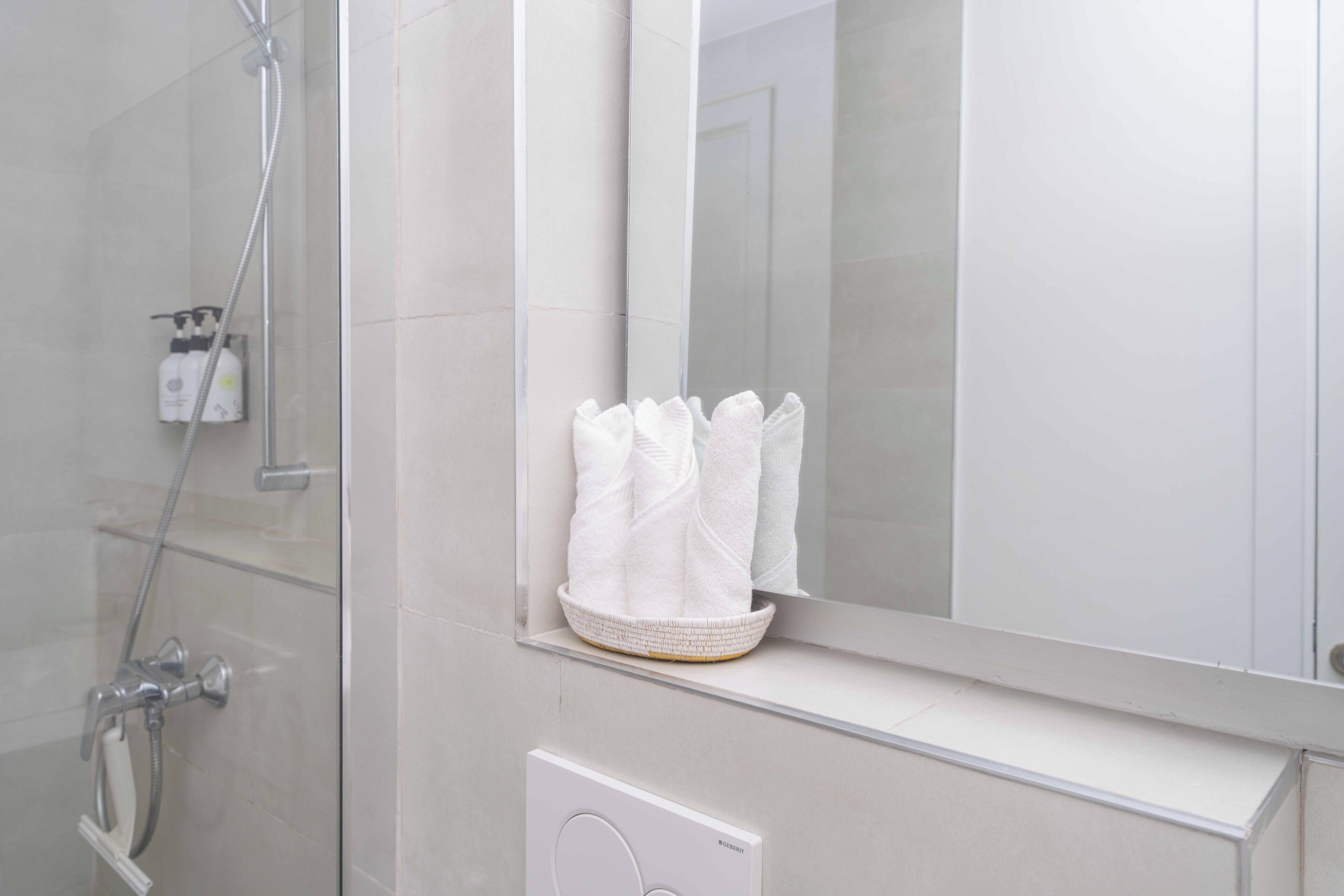 A bathroom with a glass shower enclosure, a white towel rack with folded towels, and a GEERIT brand toilet paper holder.