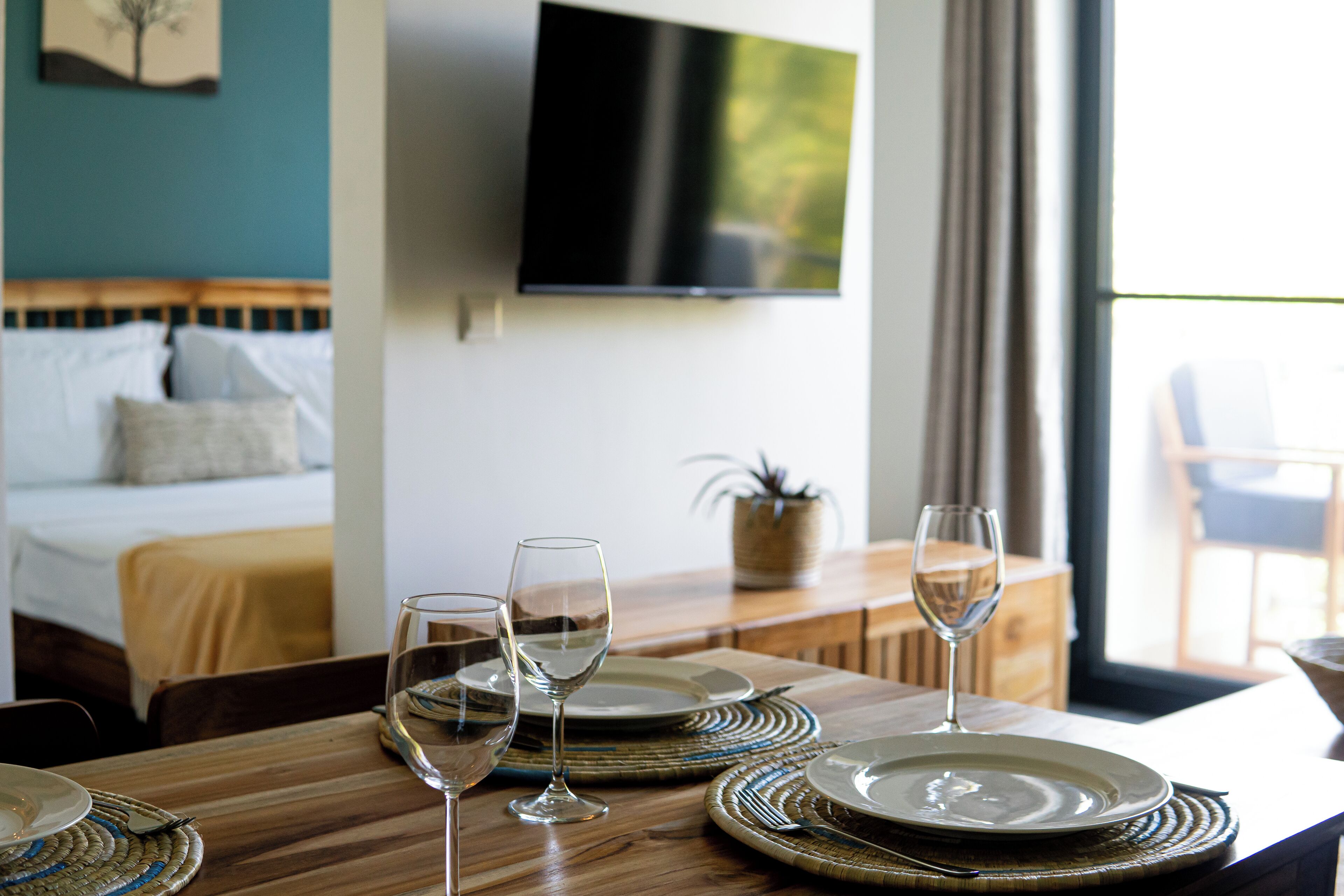 A dining table set with plates and wine glasses, a television mounted on the wall, and a bed with a headboard in the background.