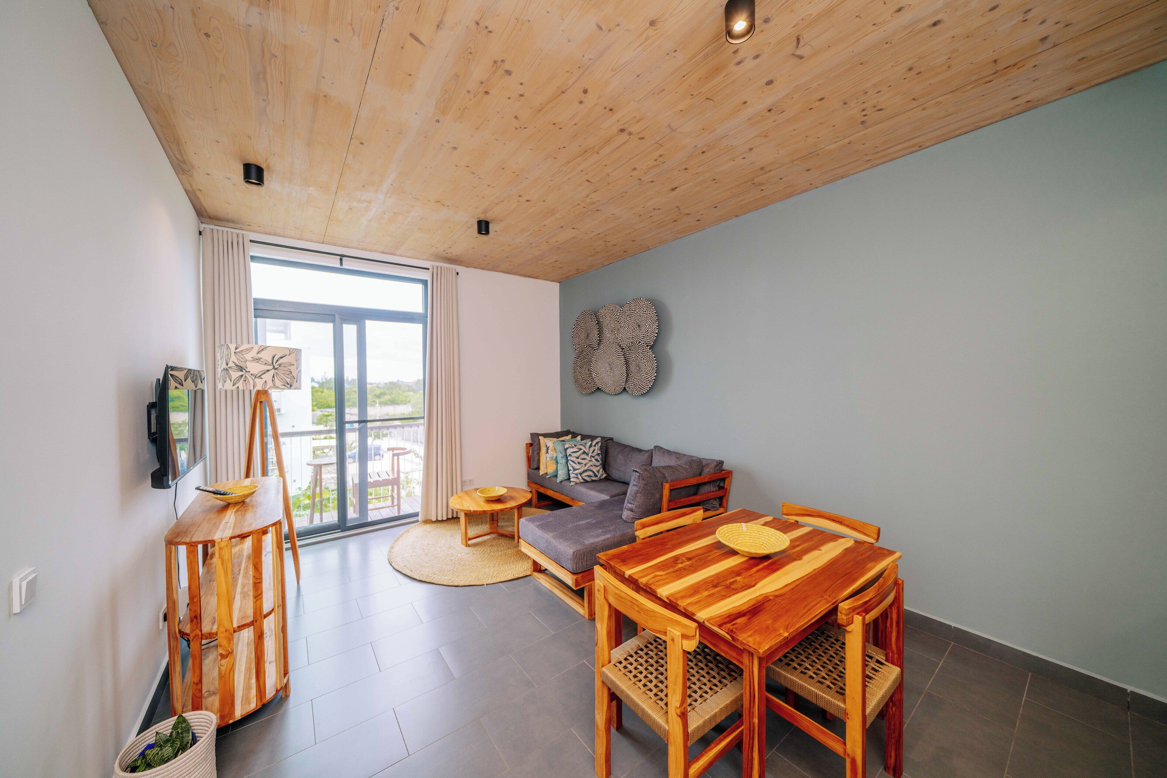 Una sala moderna con techo de madera, un sofá, una mesa de comedor con sillas, un televisor montado en la pared y un amplio ventanal con cortinas.