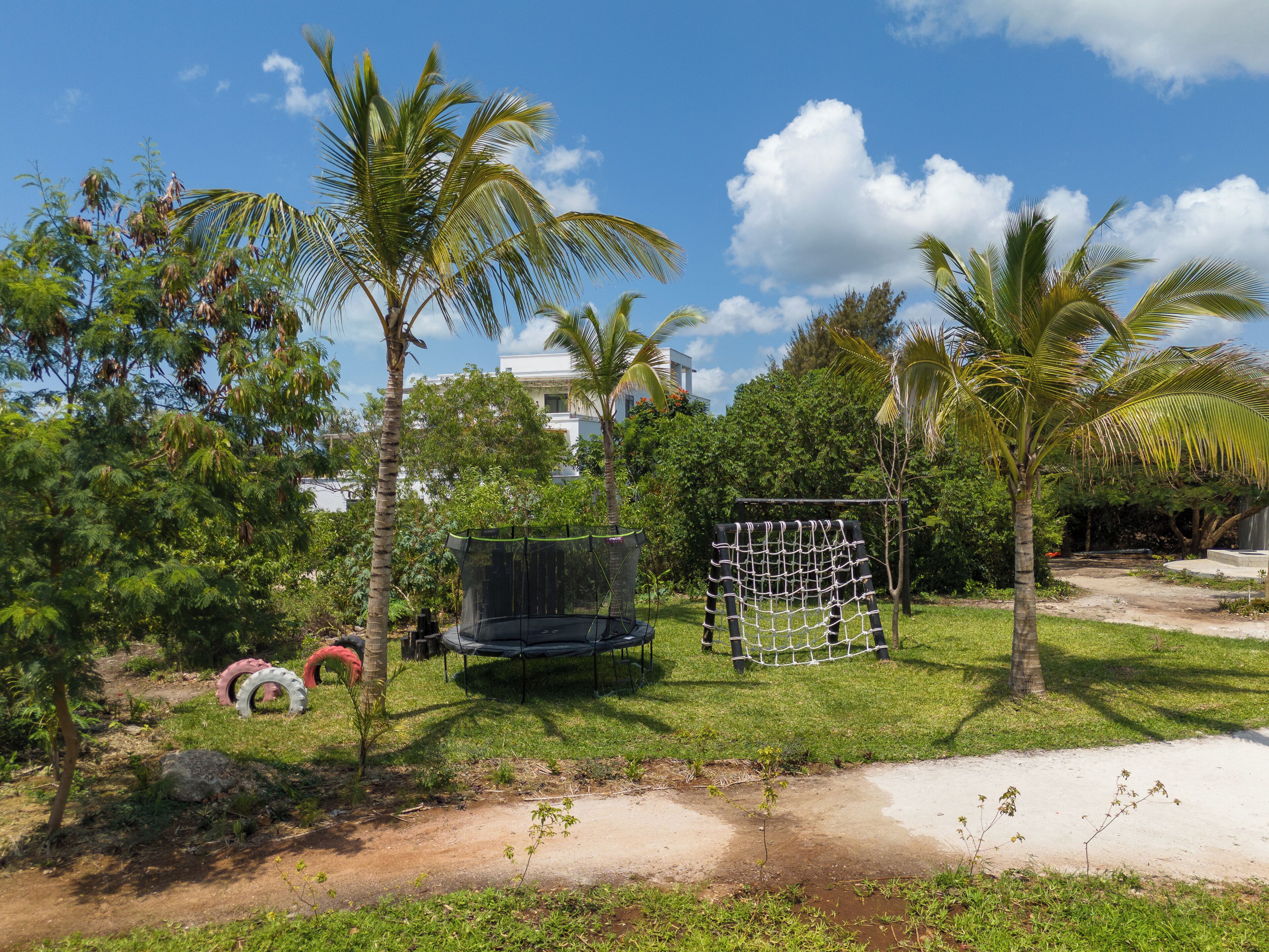 A playground with a trampoline and a netted climbing structure, surrounded by palm trees and greenery.