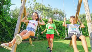 Tre bambini sull'altalena in un'area erbosa con alberi sullo sfondo.