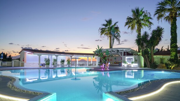 Una piscina con bordo curvo, circondata da palme e un edificio con una zona coperta sullo sfondo.
