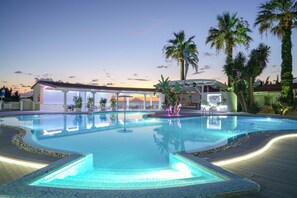 A swimming pool with a curved edge, surrounded by palm trees and a building with a covered area in the background. - Le Playe Village (Ricadi)