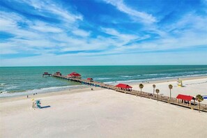Una playa con un muelle de madera que se extiende al océano, con construcciones de techo rojo a lo largo de su longitud.