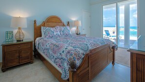 Un dormitorio con una cama de madera, dos mesitas de noche con lámparas, vista al océano a través de una puerta corrediza de vidrio y un ventilador de techo.