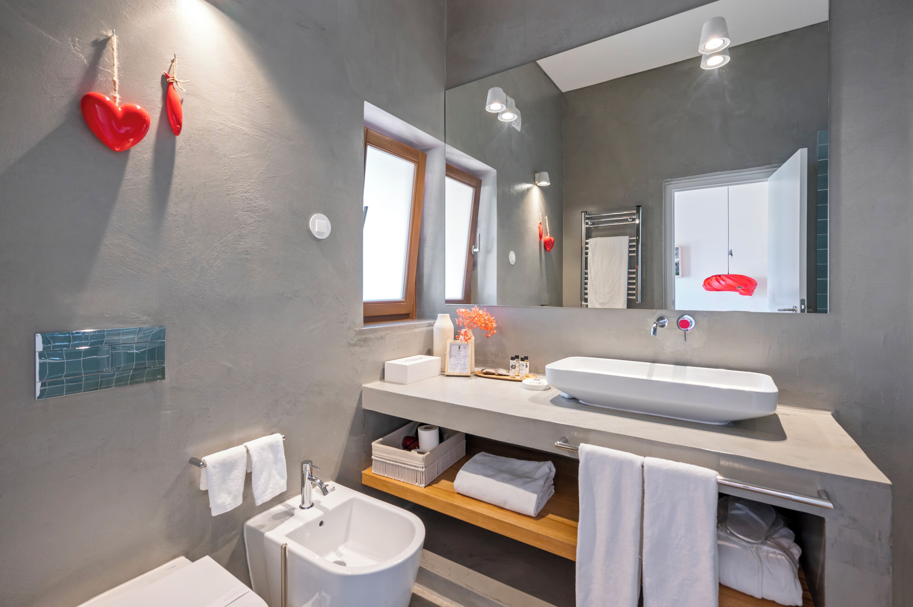 A modern bathroom with a large mirror, white sink, and a bidet. There are towels and toiletries on the counter.