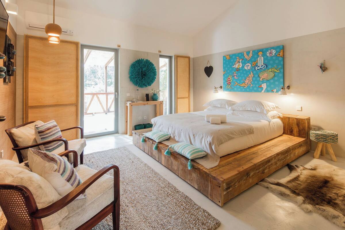 A bedroom with a wooden bed, a chair with cushions, a rug, and a wall painting.