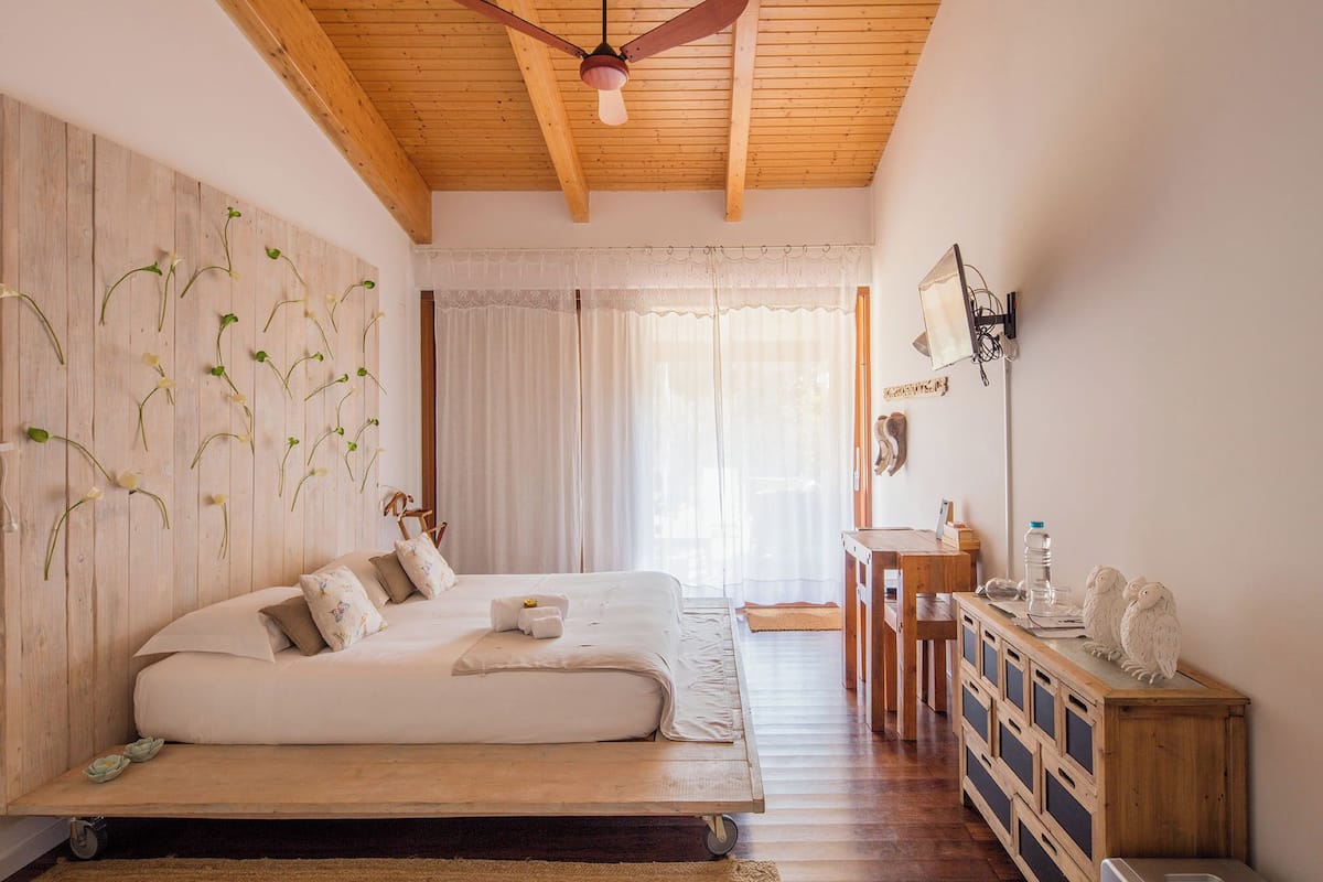 A bedroom with a wooden ceiling, a bed with white bedding, a wooden desk, and a wall-mounted TV.