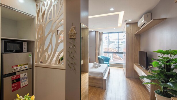 Una habitación de hotel moderna con kitchenette, una cama con ropa blanca, una silla azul, un ventanal grande y una planta en maceta.