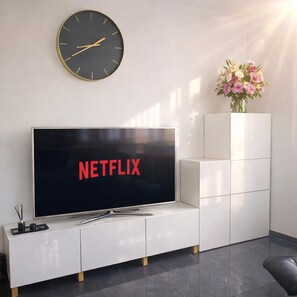 Una sala moderna con un televisor de pantalla plana mostrando 'NETFLIX', un mueble blanco para equipo electrónico, un reloj negro y un florero con flores.