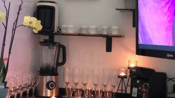 Zona de bar con una máquina de café, vasos y velas. Hay un televisor en la pared y un cuadro enmarcado encima.