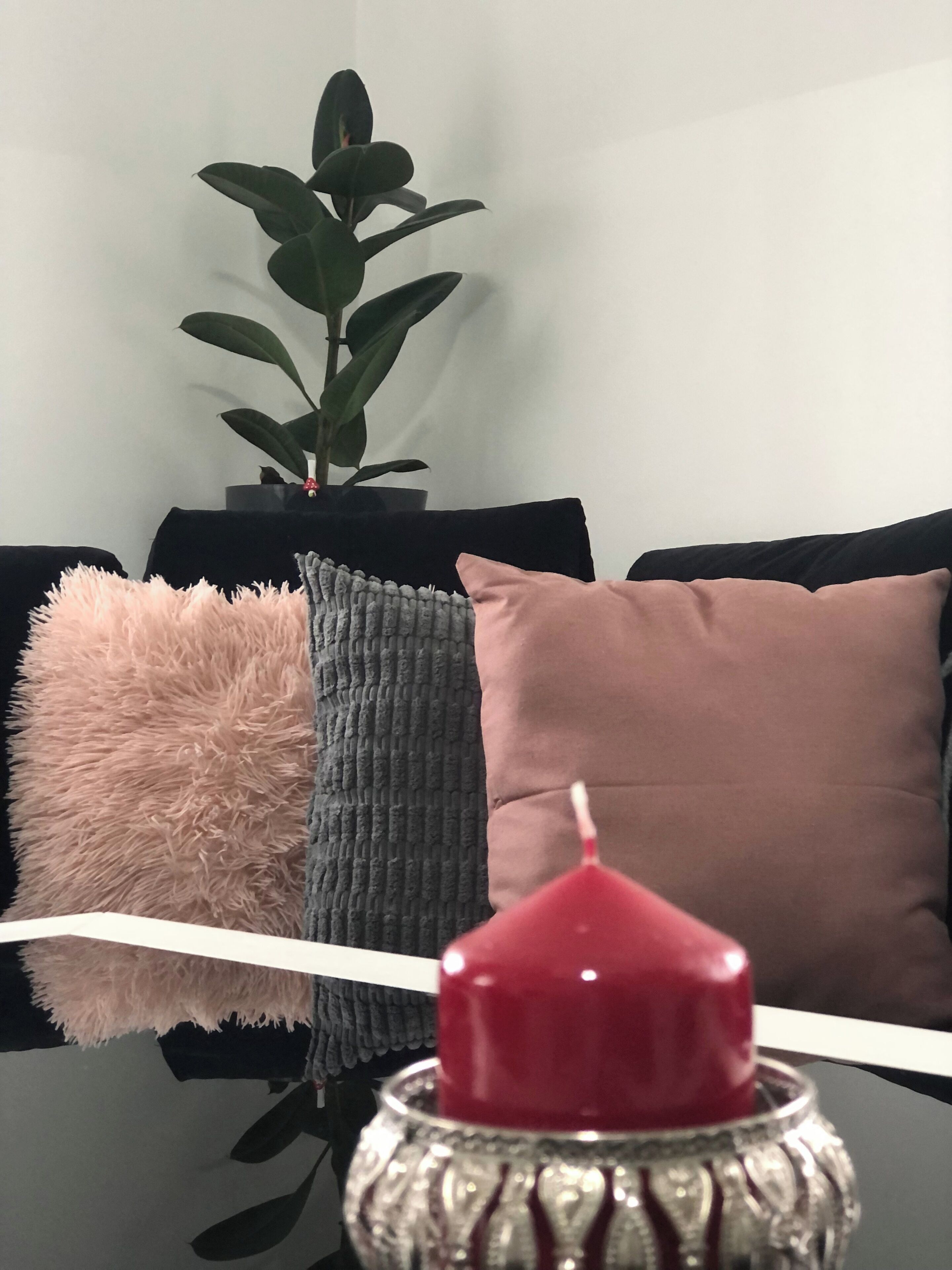 A black table with a red candle in a decorative holder, a fluffy pink pillow, and a grey knitted pillow on a black sofa.
