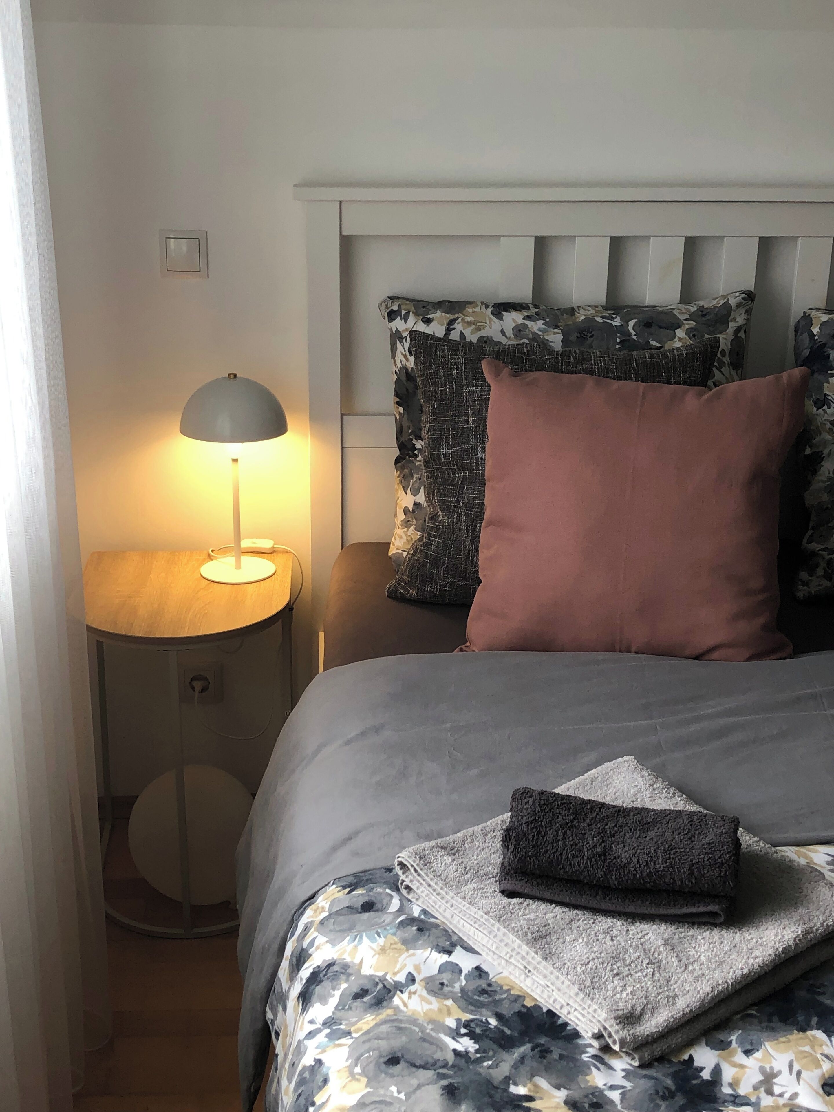 Una cama bien hecha con ropa de cama de estampado floral, una lámpara de noche y una mesita redonda con un teléfono.