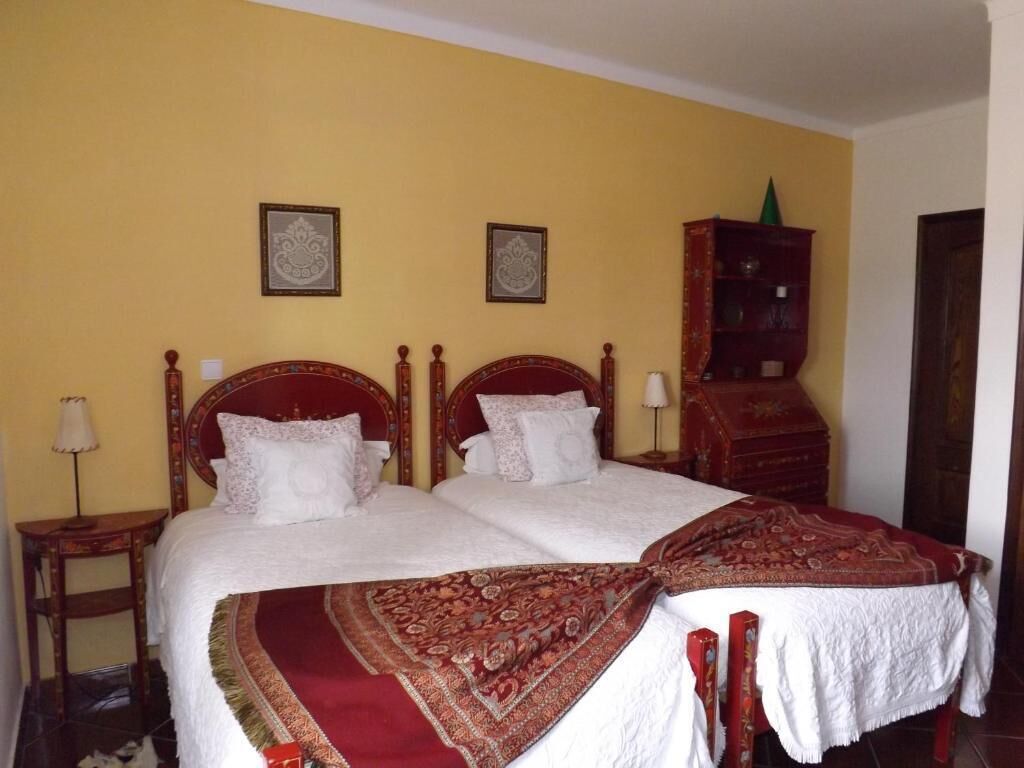Una cama doble con colcha roja y blanca, dos mesitas de noche, un armario de madera y dos cuadros en la pared.