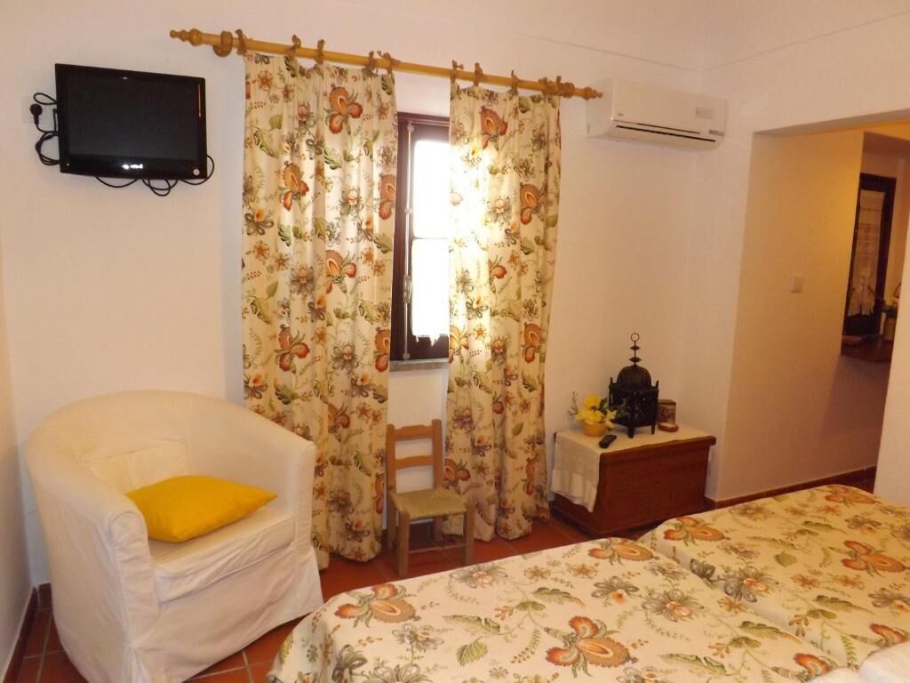 A bedroom with a bed, a chair with a yellow cushion, a small table with a lamp, and a television mounted on the wall.