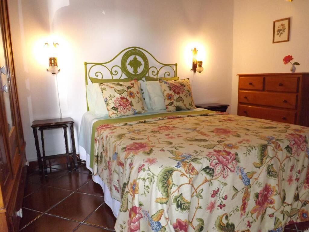 Un dormitorio con colcha estampada, cabecero de metal verde, dos lámparas de noche, cómoda de madera y una mesita.