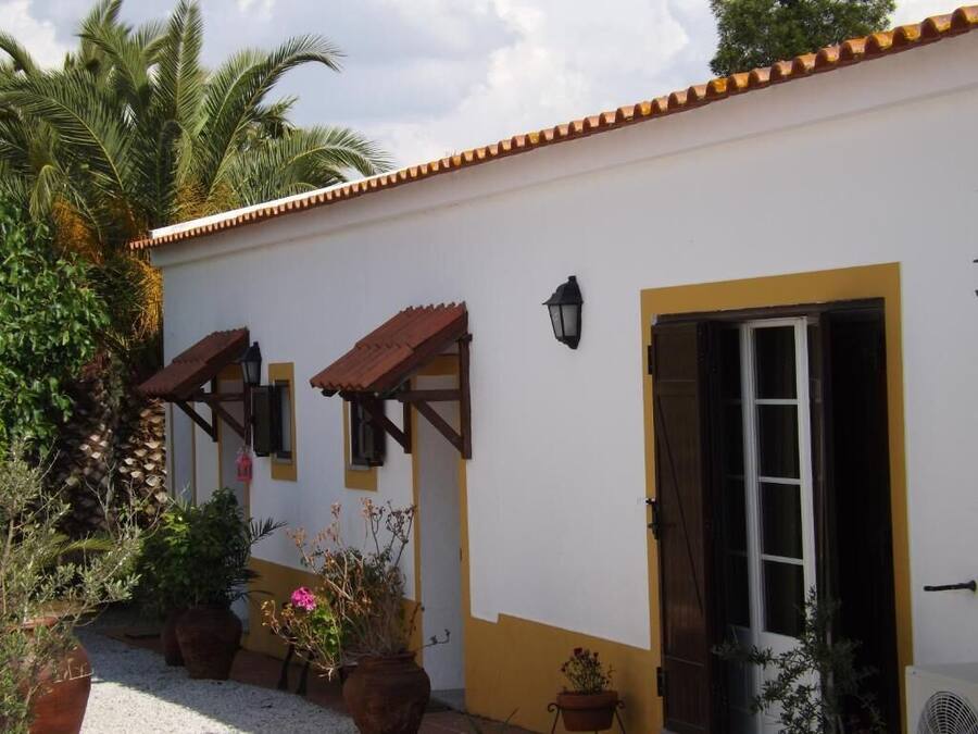 Un edificio blanco con detalles amarillos y techo de tejas. Se observan dos ventanas con contraventanas de madera y una puerta.