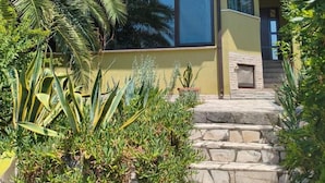 Una casa con una escalera de piedra que lleva a una puerta, rodeada de exuberante follaje y una montaña al fondo.