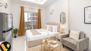 A modern hotel room with a bed, a sofa, a dining table, a mirror, and a window with curtains. - Vogue | Hera Tower Sports City near First Avenue Mall (Dubai)
