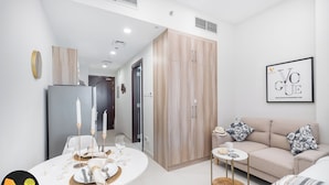 A modern living room with a sofa, a dining table set for two, and a kitchen area with a refrigerator. - Vogue | Hera Tower Sports City near First Avenue Mall (Dubai)