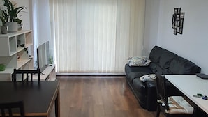 A living room with a black sofa, a white table, and a TV on a stand. There are potted plants on a shelf and a window with vertical blinds. - 1 Bed Apartment Sleeps 2 With Wi-fi & Tv (London)