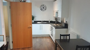 A compact kitchen with white cabinets, a wooden door, and a dining area with a table and chairs. - 1 Bed Apartment Sleeps 2 With Wi-fi & Tv (London)