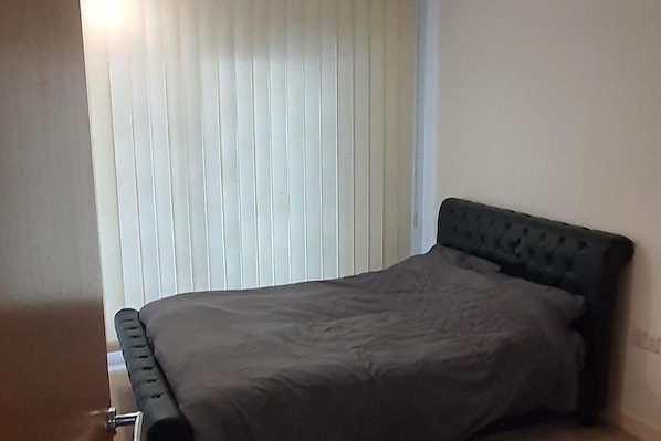 Una cama individual con cabecera oscura en una habitación con ventana cubierta por persianas verticales.