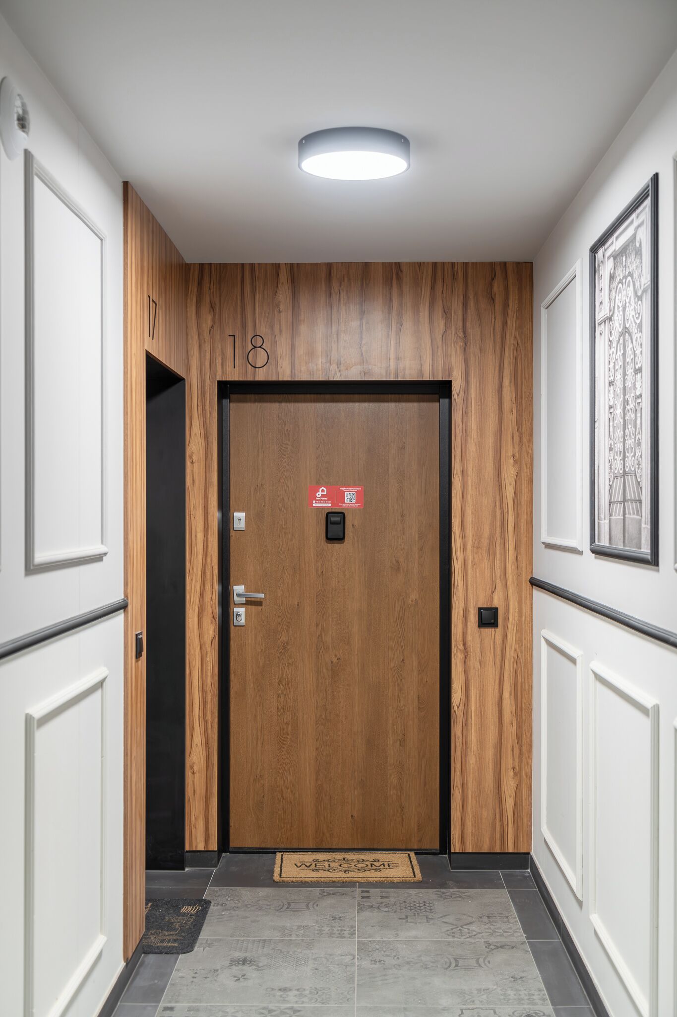 Un pasillo con una puerta de madera marcada con el número 18, una alfombra de bienvenida y un cuadro enmarcado en la pared.
