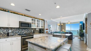 A kitchen with white cabinets, a large island, a microwave, and a stove. There is a dining area with a table and chairs. - Majestic Beach Towers 2-404 (Panama City Beach)