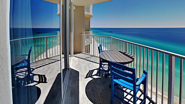 A balcony with a table and chairs, overlooking a clear blue sea and a pier.