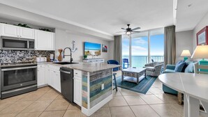 A modern kitchen with white cabinets, a built-in oven, and a dishwasher. There is a dining area with a table and chairs, and a living room with a sofa and a view of the ocean.