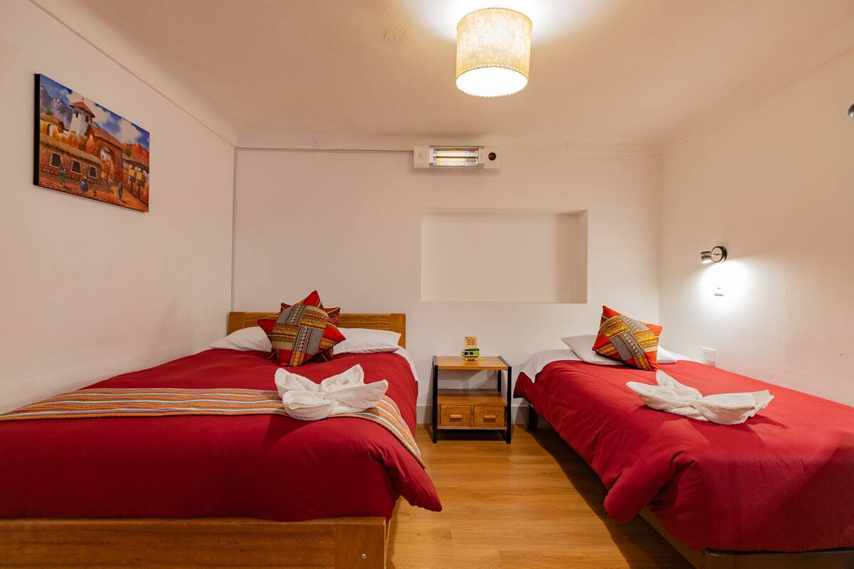 Ein Hotelzimmer mit zwei Betten, jeweils mit roter Bettwäsche und dekorativen Kissen, einem Nachttisch aus Holz mit einer Lampe sowie einem Bild an der Wand.