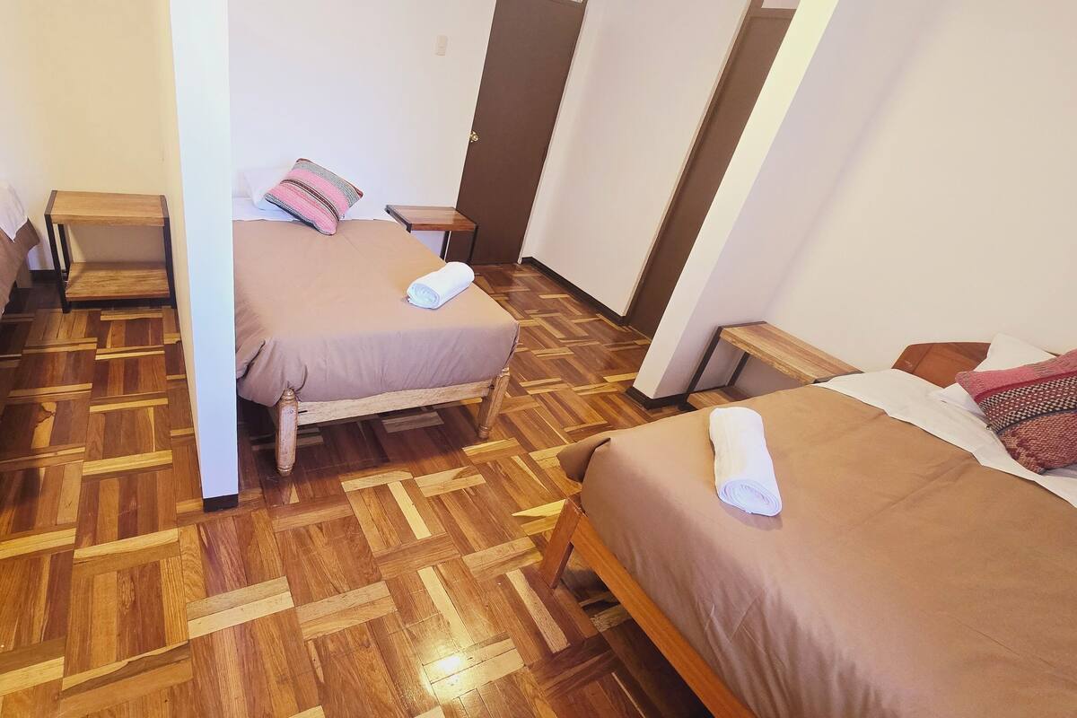 Ein Hotelzimmer mit drei hölzernen Betten, jedes mit einer braunen Bettdecke und einem aufgerollten weißen Handtuch. Das Zimmer verfügt über einen Parkettfußboden und eine Holztür.