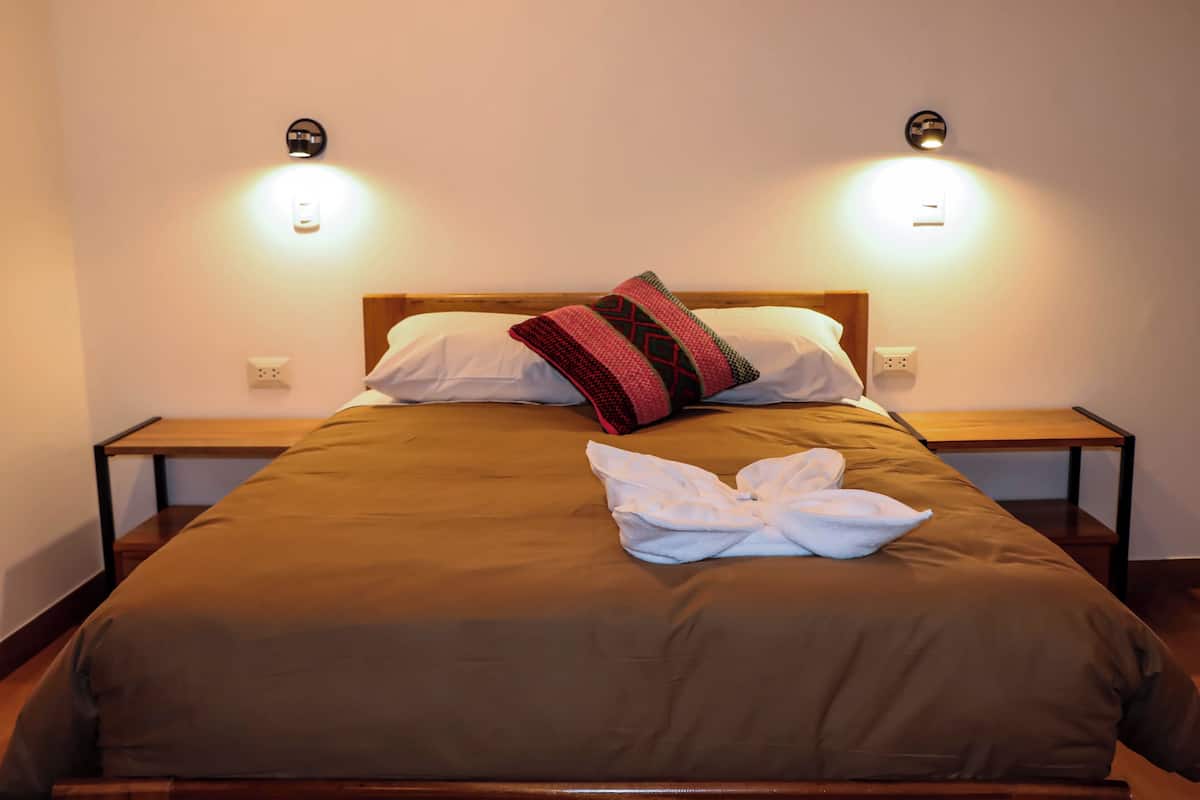 Ein ordentlich bezogenes Bett mit einer braunen Tagesdecke, einem weißen Kopfkissen und einem Zierkissen. An beiden Seiten des Bettes stehen Nachttische mit Lampen.