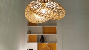 Une pièce avec un luminaire suspendu en fibres tressées, des murs blancs et une poutre en bois au plafond. On y trouve des étagères remplies de divers objets et un portant à vêtements suspendu.