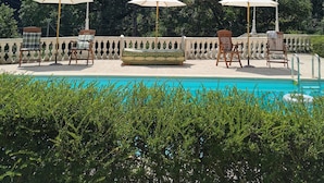 Un espace piscine agrémenté de trois chaises en bois et de deux parasols blancs, entouré d’une végétation luxuriante et surplombé d’un ciel dégagé.