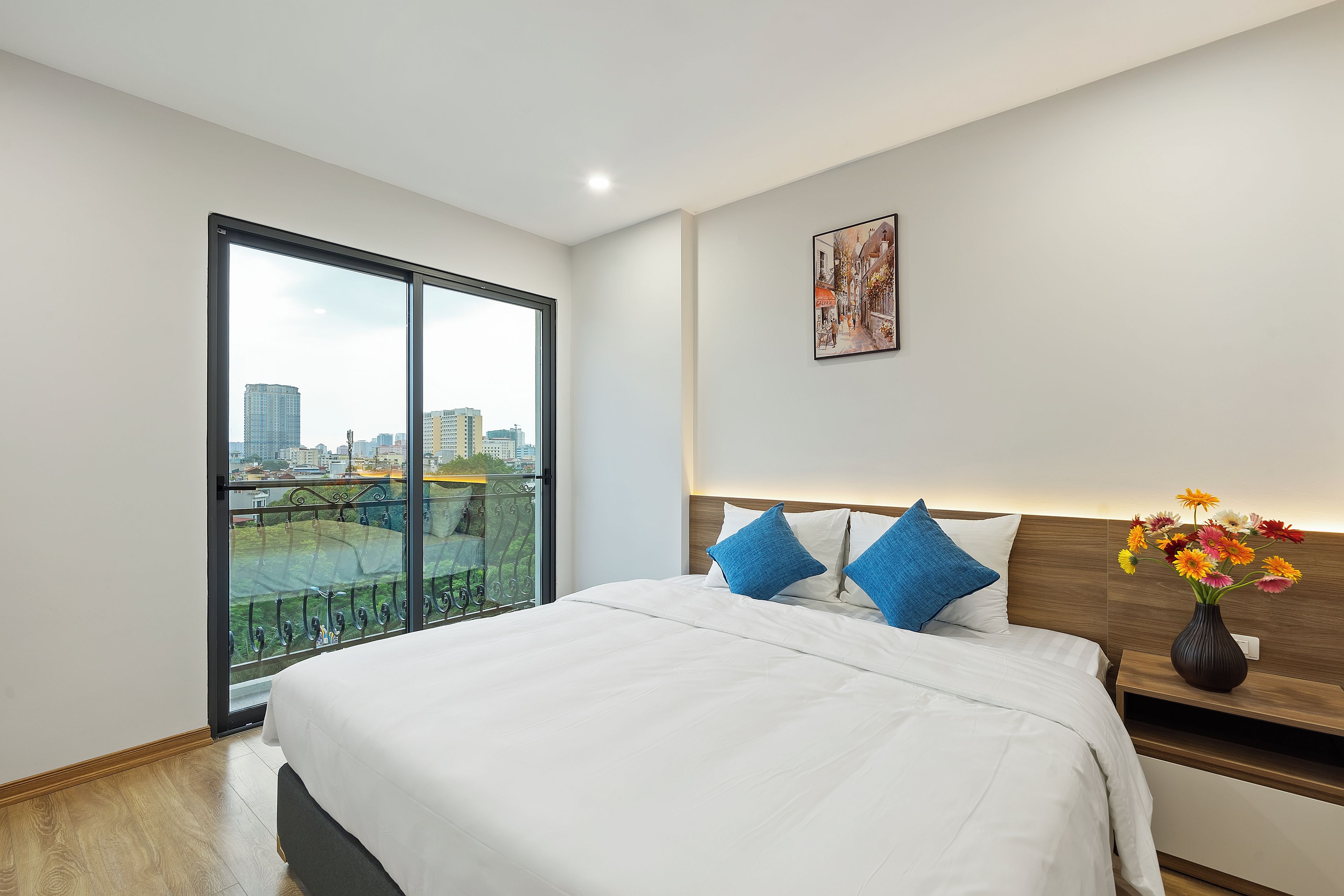 A modern hotel room with a large bed, a nightstand with a vase of flowers, a city view through a large window, and a framed picture on the wall.