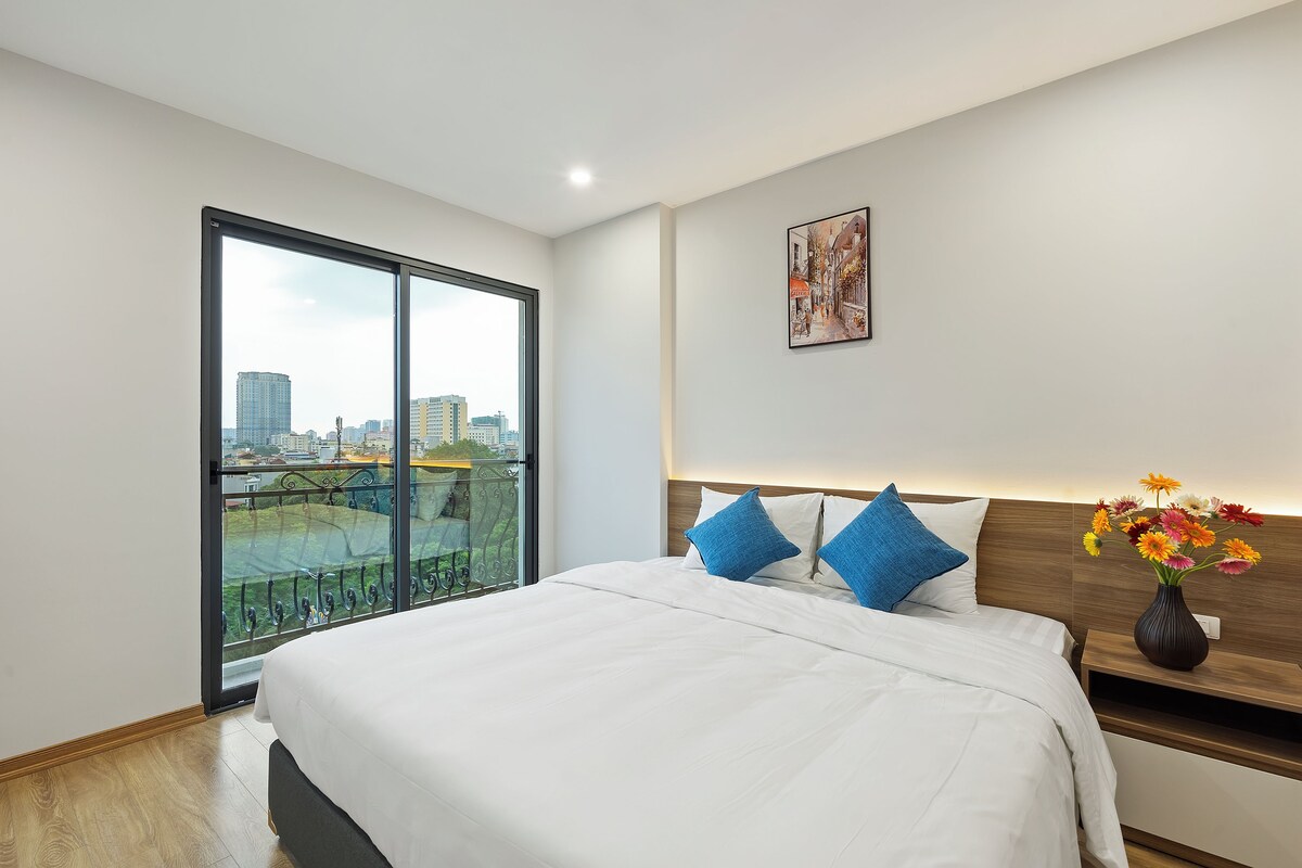 A modern hotel room with a large bed, a nightstand with a vase of flowers, a city view through a large window, and a framed picture on the wall.