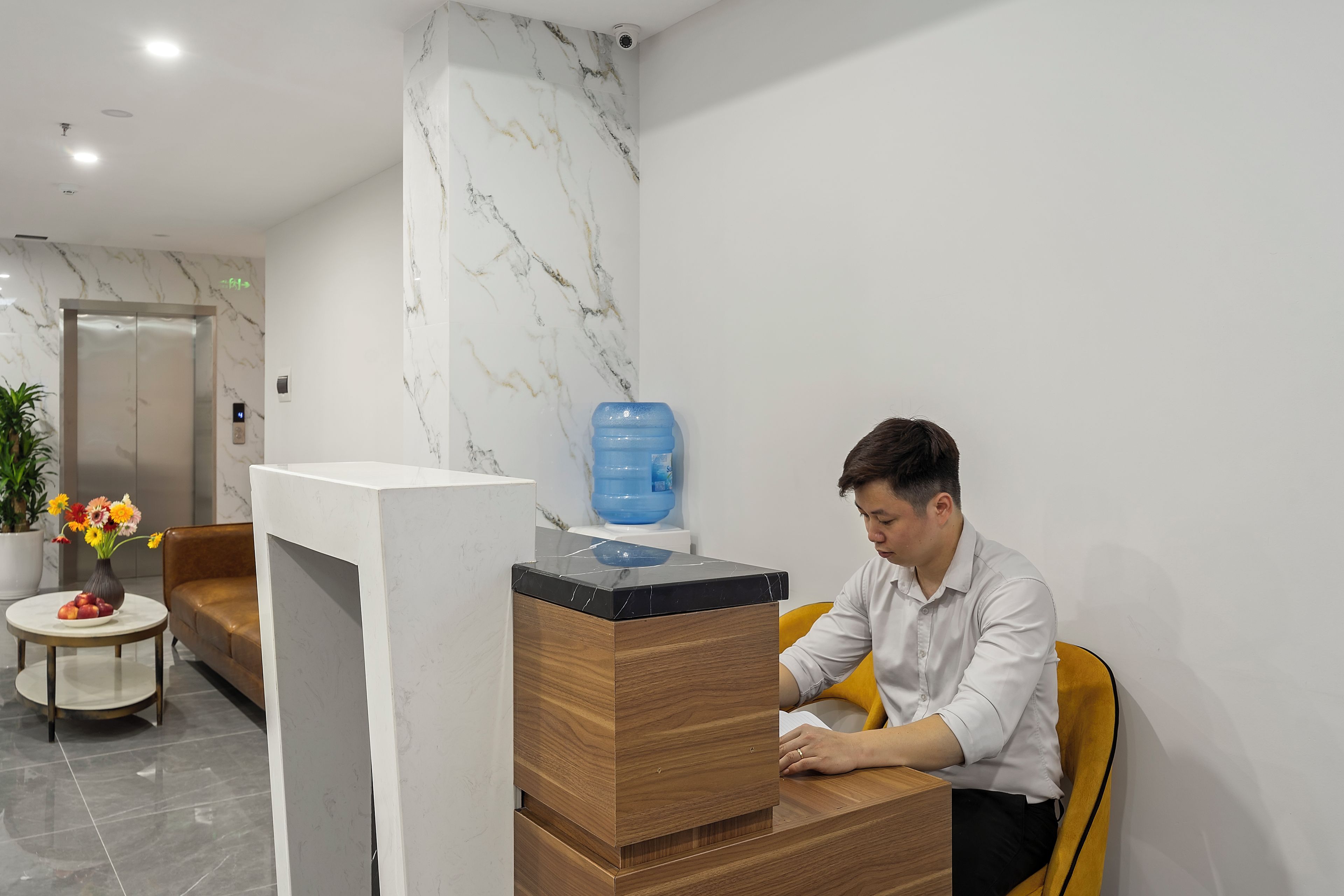 Un mostrador de recepción con pared de mármol, dispensador de agua y una zona de estar con sofá y una mesita con flores.