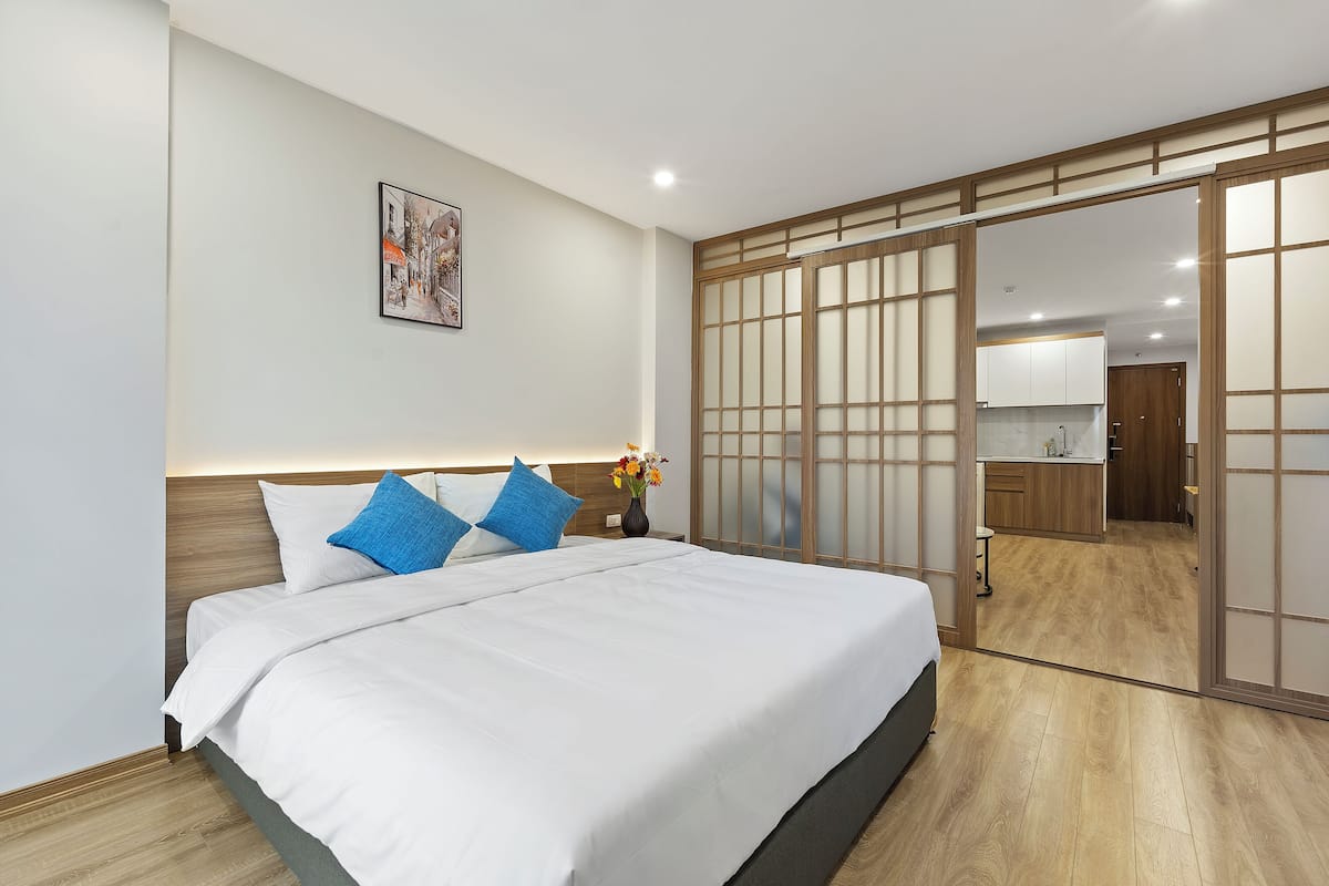 A modern hotel room with a large bed, a wooden headboard, a nightstand with a vase of flowers, and a sliding door leading to a kitchen area.