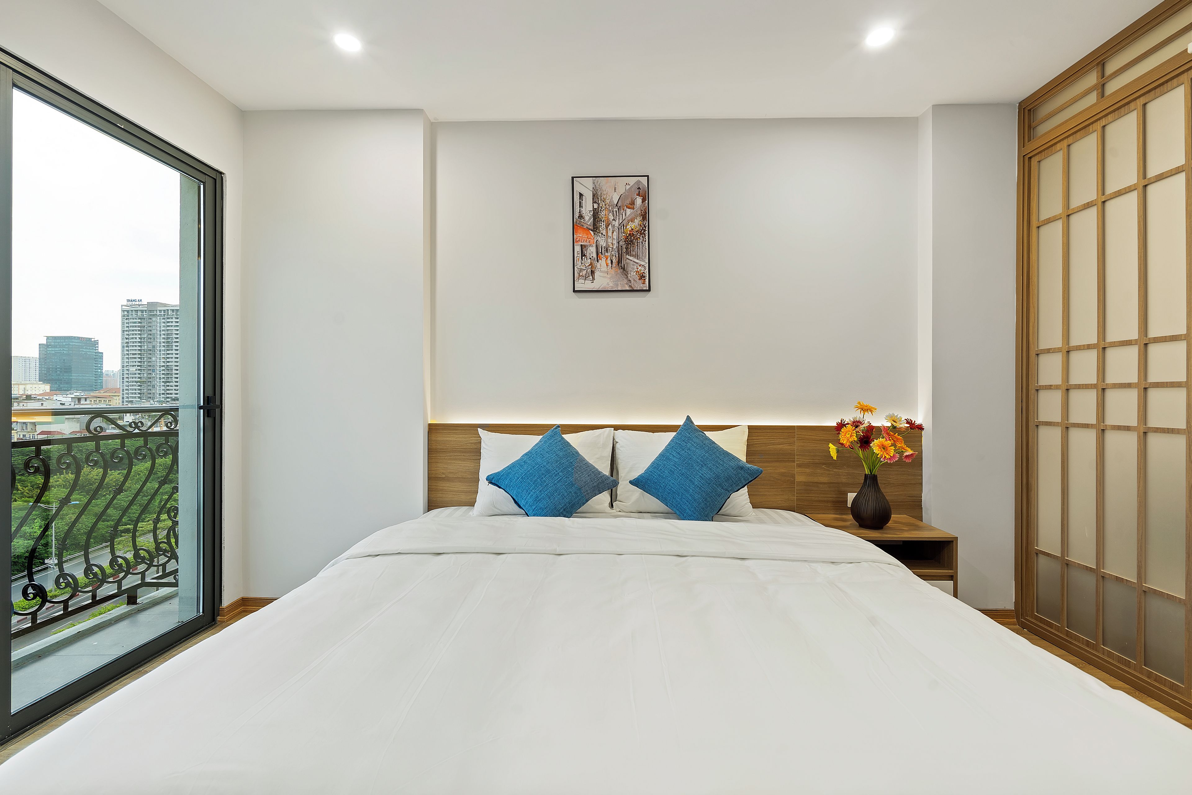 A hotel room with a large bed, a nightstand with a vase of flowers, a painting on the wall, and a view of the city through a window.