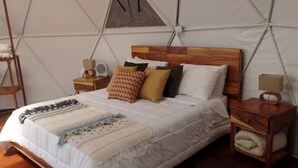 Un lit avec une tête de lit en bois, une couette blanche et plusieurs oreillers. On y trouve une table de chevet en bois avec une lampe et un ventilateur. Le plafond de la pièce a la forme d’une structure semblable à une tente.