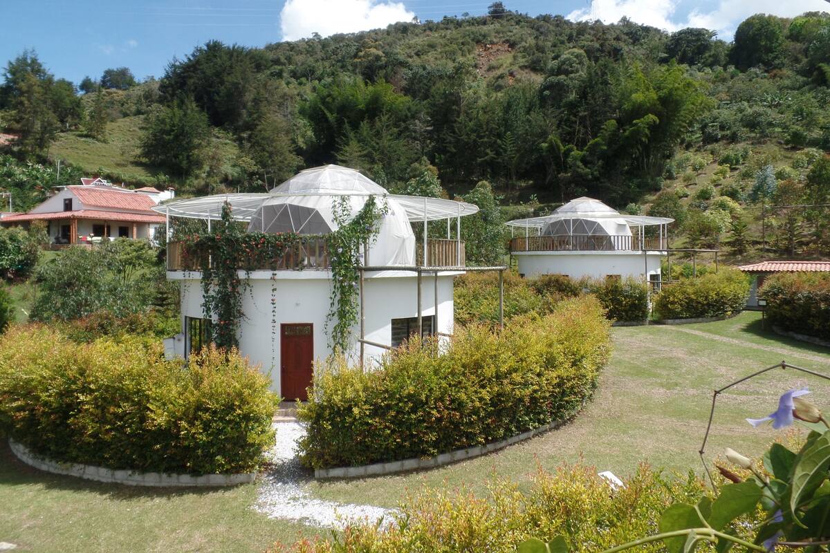 A white building with a dome structure, surrounded by lush greenery and a hill in the background.