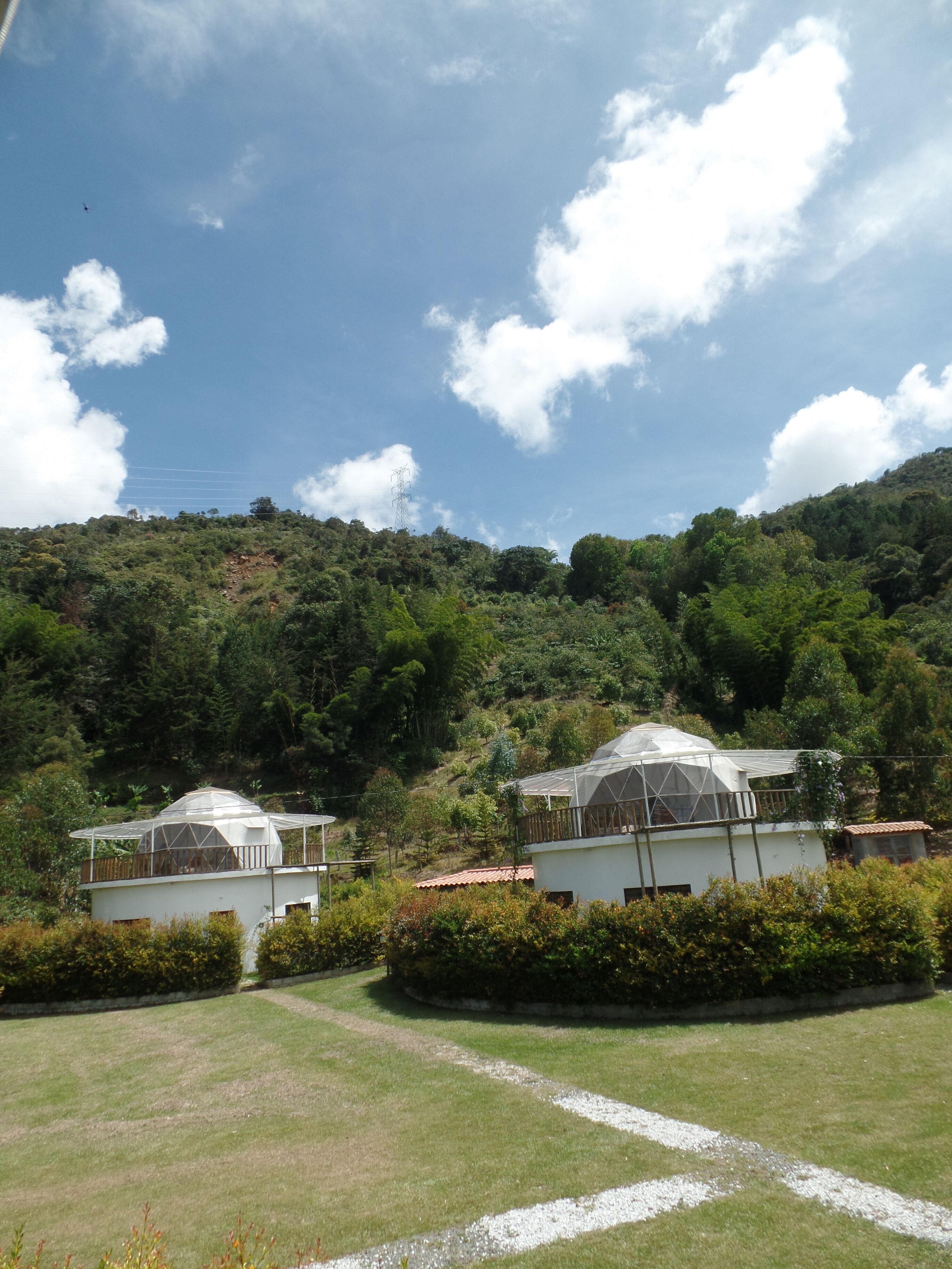 A hillside property with two white buildings, each with a large covered patio. The property features a well-maintained lawn and a gravel path leading to the buildings.