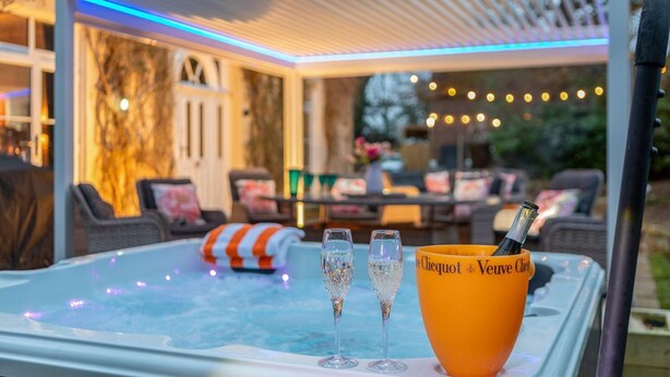 A hot tub with three glasses and a bottle of champagne, surrounded by outdoor seating and string lights.