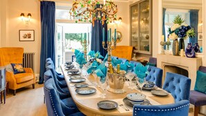 A formal dining room with a long table set for a meal, blue upholstered chairs, a chandelier, and a view of the outdoors through a large window.