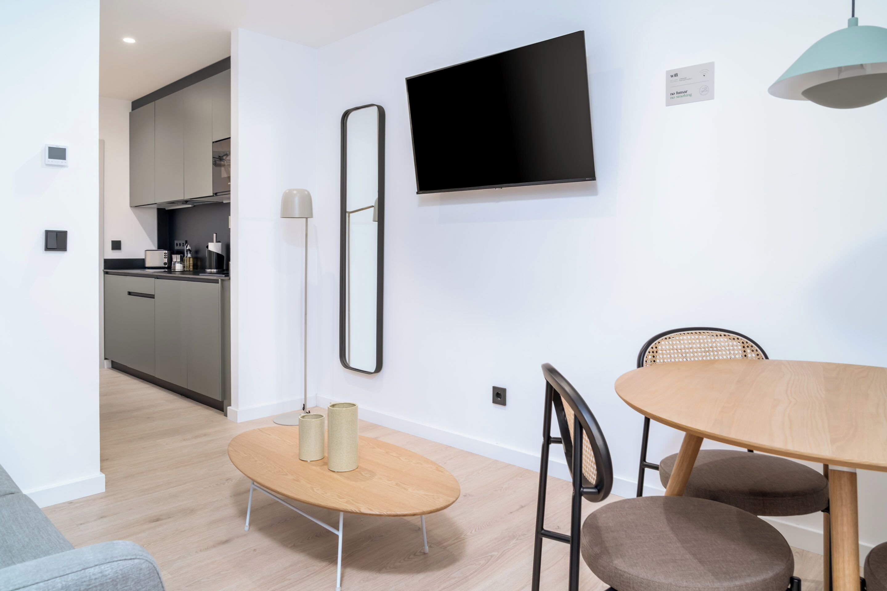 Una cocina moderna con armarios grises, un televisor de pantalla plana montado en la pared y un comedor con mesa y sillas de madera.